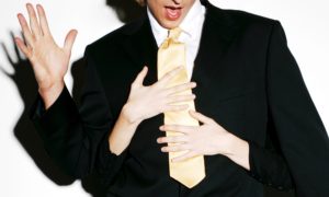 A man in a black suit, white shirt and yellow tie looking comically surprised at what look to be a woman's hands wrapped around him groping him from behind.