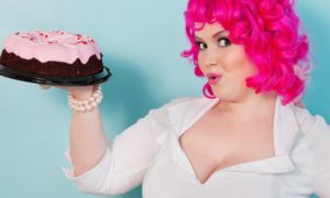A cheeky pretty woman with hot pink hair holding a pink-frosted chocolate cake.